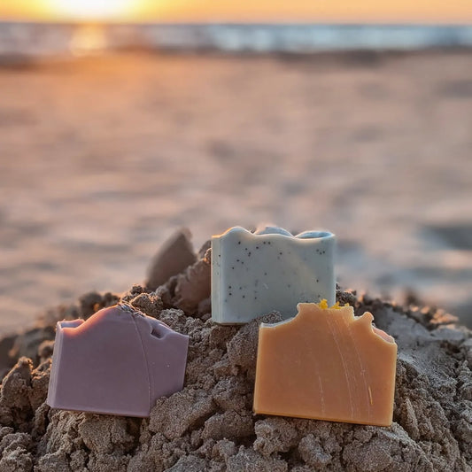 Three colorful soap bars on a sandy beach with a blurred ocean background