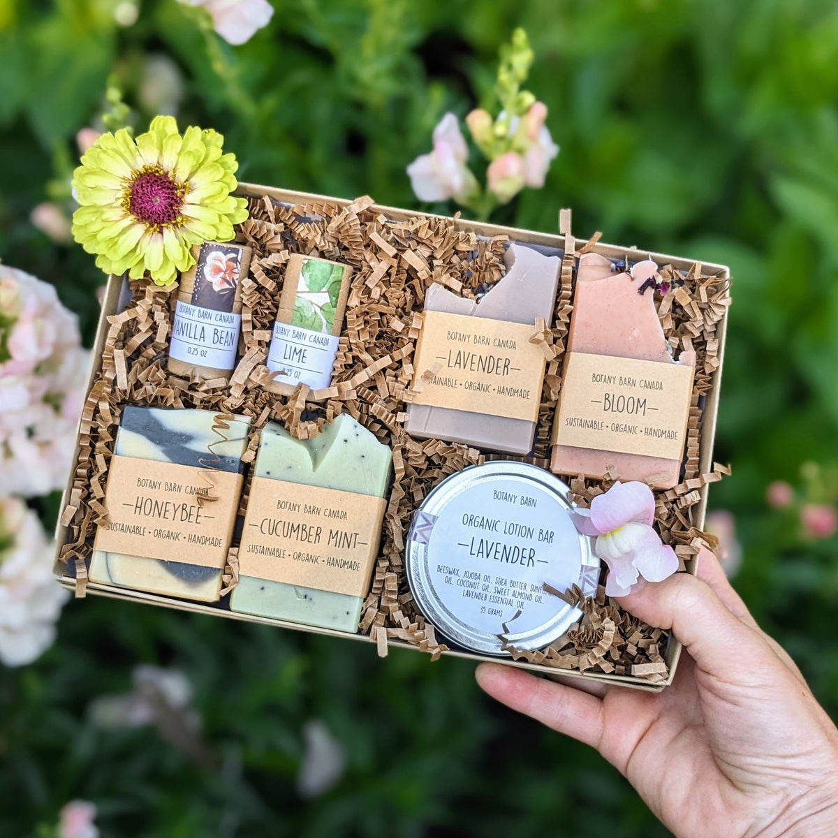 A gift set of handmade soaps, organic lotion in a metal tin, and eco-friendly lip balm tubes, all packaged in a kraft gift box with decorative flowers on top.