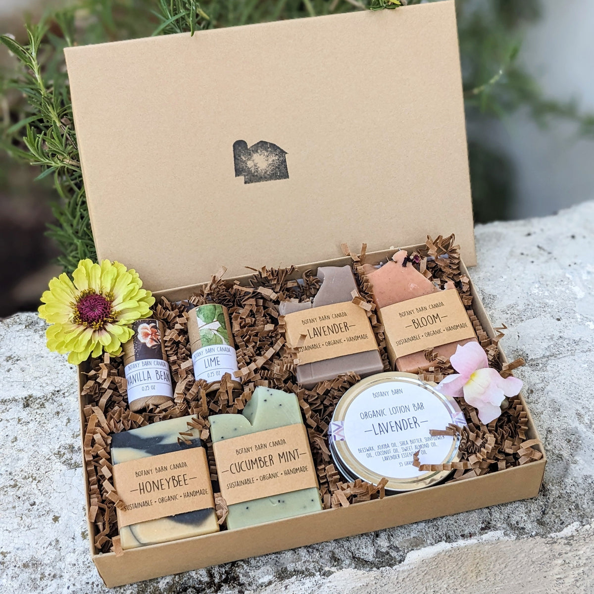 A gift set of handmade soaps, organic lotion in a metal tin, and eco-friendly lip balm tubes, all packaged in a kraft gift box with decorative flowers on top.