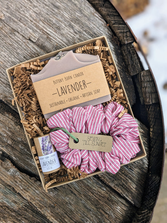 An eco-friendly gift set presented in a kraft gift box, containing a lavender-colored soap bar, a pink and white striped handmade scrunchie, and a lip balm tube.