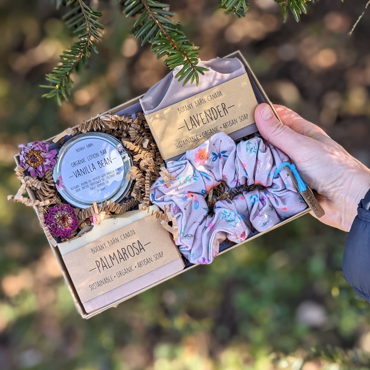 A hand holding a kraft gift box with a selection of eco-friendly beauty products including two soap bars, a lotion bar in a metal tin, and a handmade scrunchie with a floral pattern.