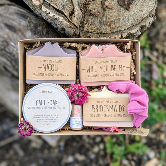 A gift set including soap bars, bath salts, lip balm and scrunchie. Custom soap labels read: Nicole, will you be my bridesmaid?