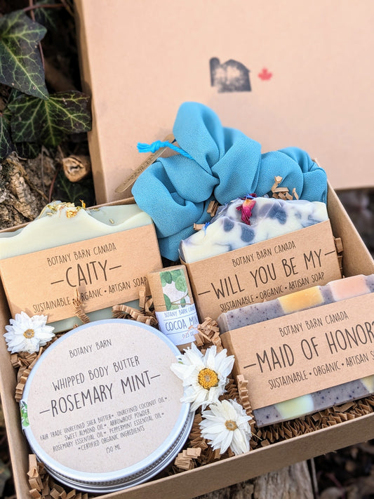 A gift set including handmade soaps, body butter, lip balm, and a scrunchie, packaged in a kraft box with custom labels and tied with a bow.