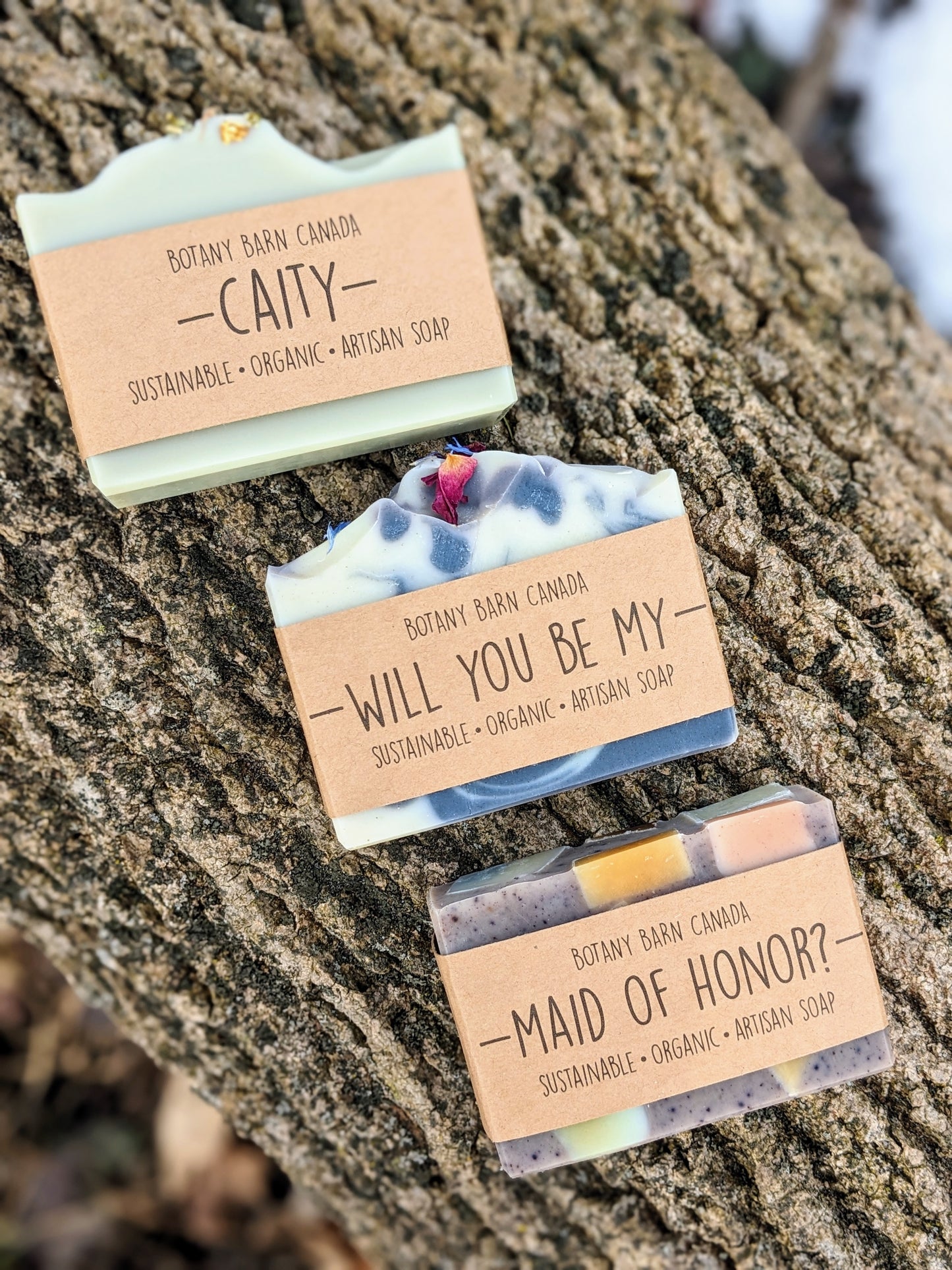 Three soap bars with custom labels that read "Caity, will you be my maid of honor?"