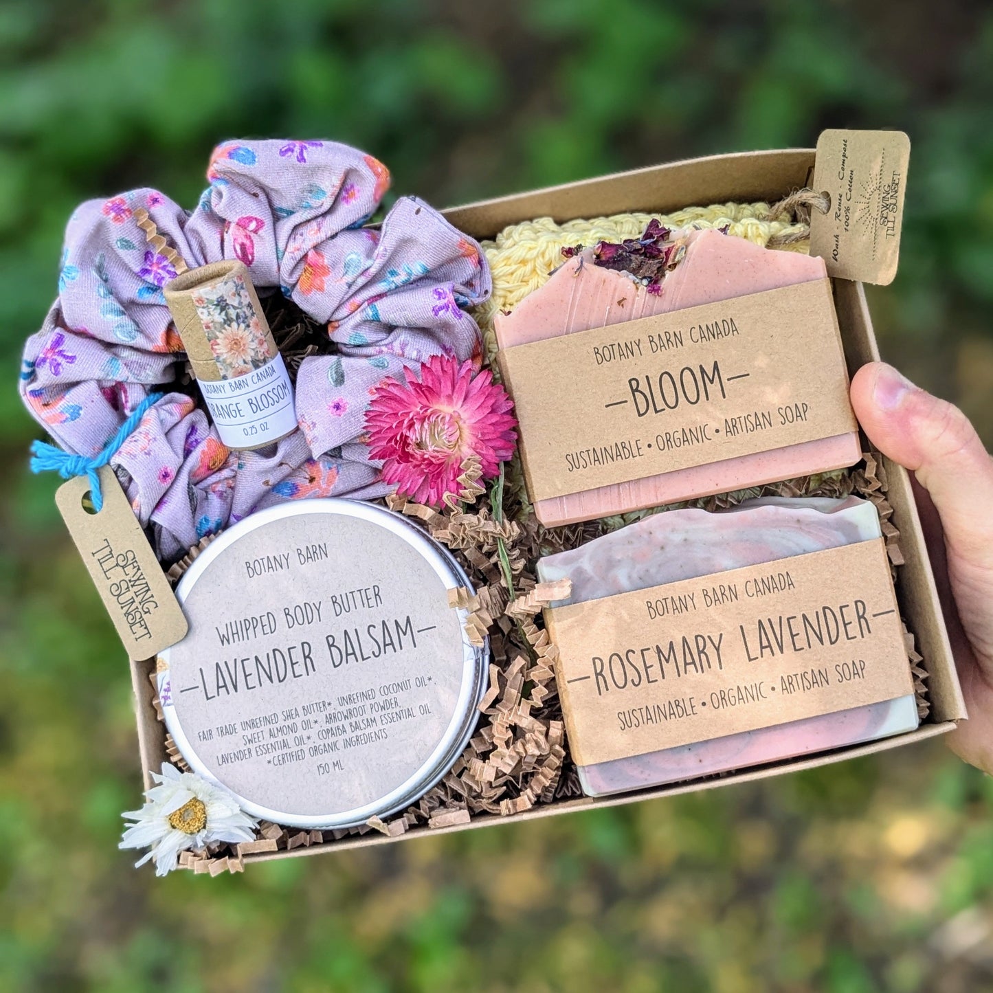 A floral spa gift set displayed on a wooden surface, including two soap bars, a crocheted washcloth, a metal tin with whipped body butter, a handmade scrunchie, and a lip balm tube, all arranged in an open gift box.