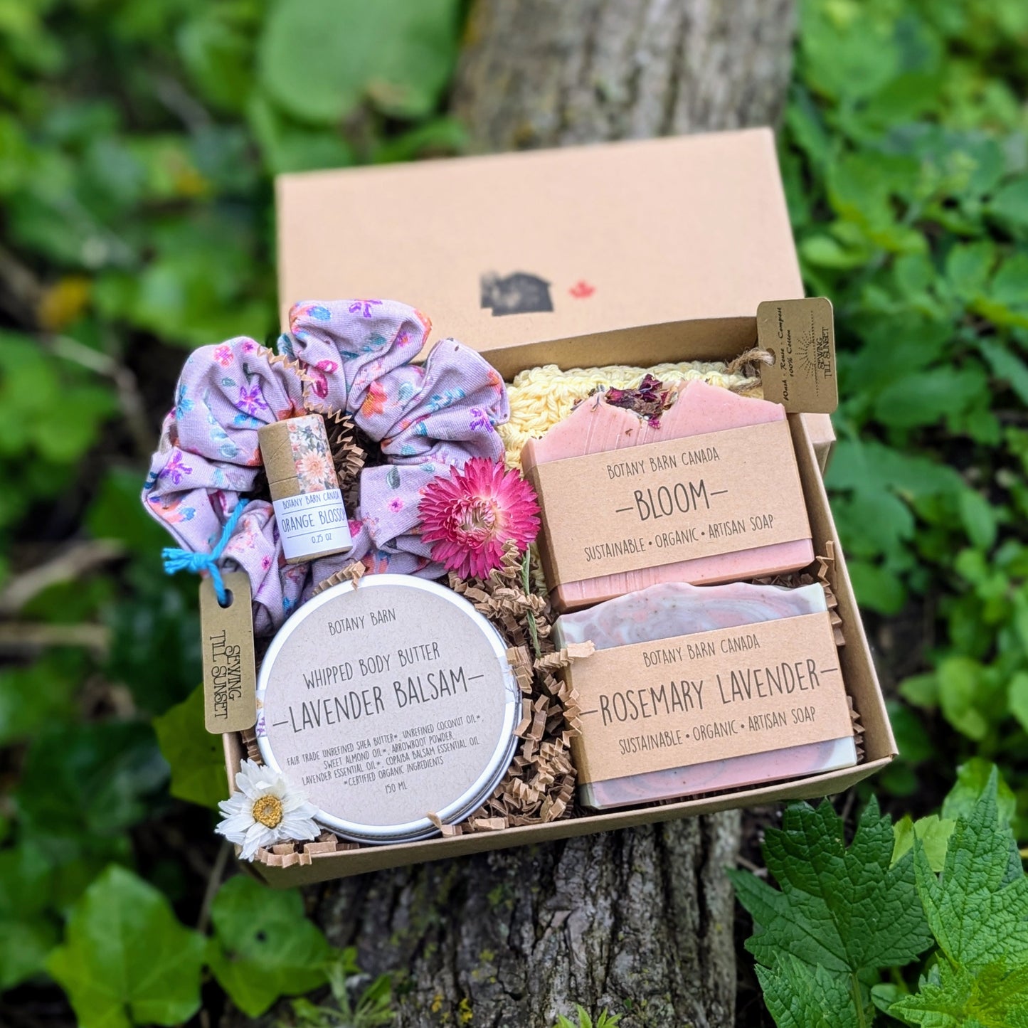 A floral spa gift set displayed on a wooden surface, including two soap bars, a crocheted washcloth, a metal tin with whipped body butter, a handmade scrunchie, and a lip balm tube, all arranged in an open gift box.