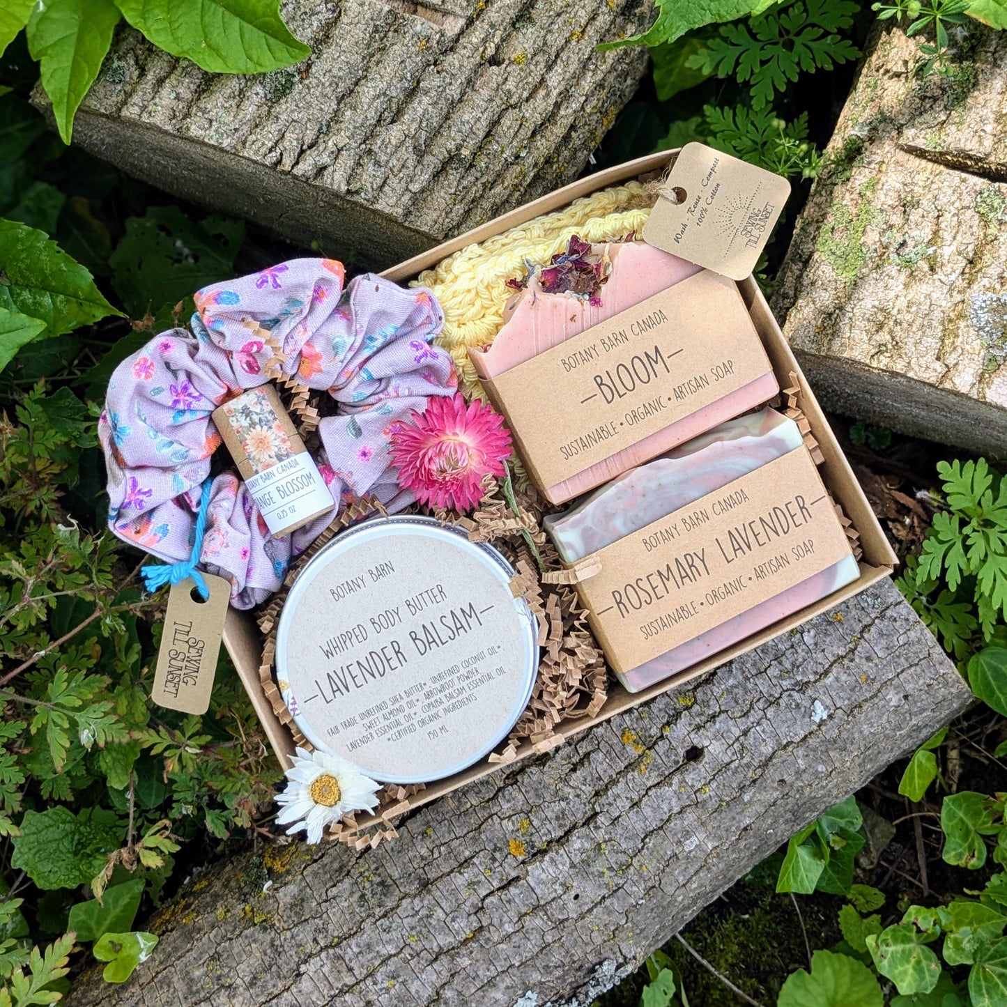 A floral spa gift set displayed on a wooden surface, including two soap bars, a crocheted washcloth, a metal tin with whipped body butter, a handmade scrunchie, and a lip balm tube, all arranged in an open gift box.