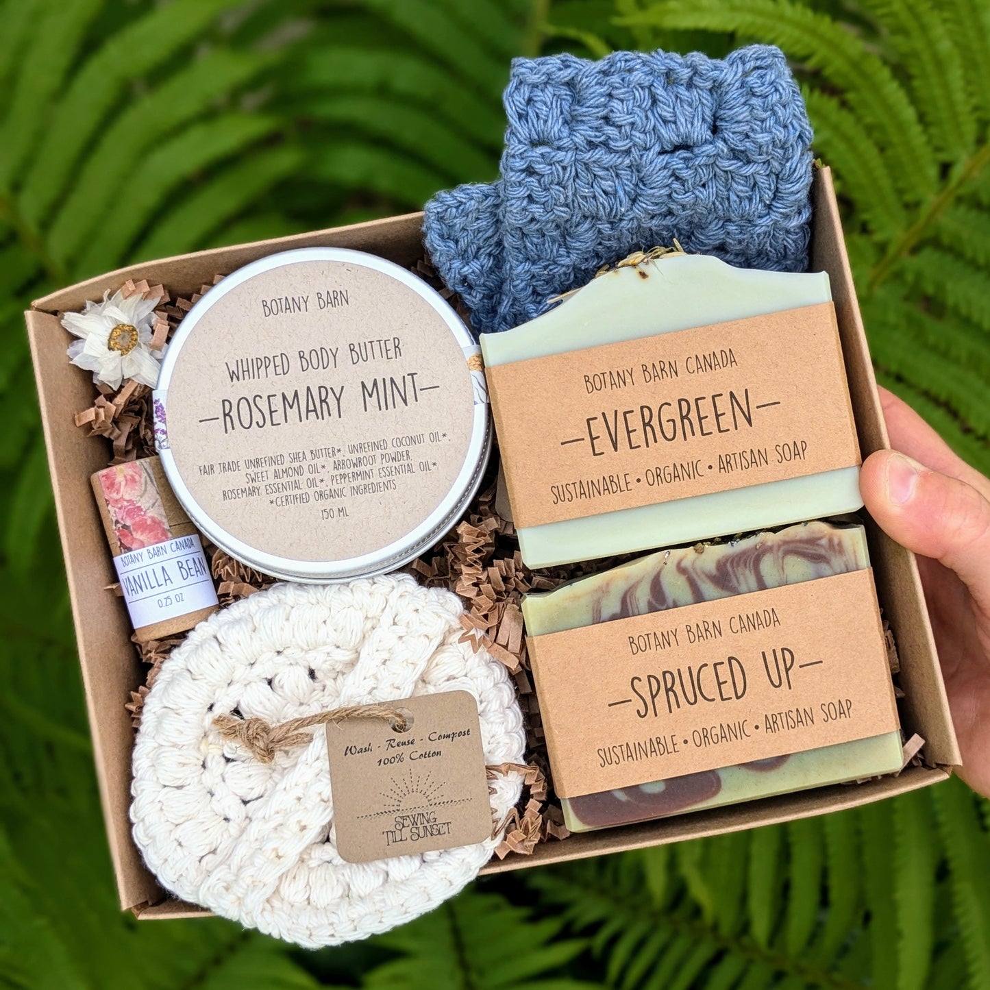 A gift set box containing two soap bars, a blue crocheted washcloth, a metal tin of body butter, a round scrubbie, and a tube of lip balm.