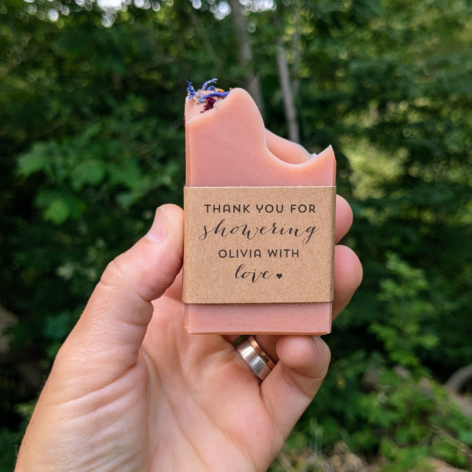A pink handmade soap bar with custom labels reading "thank you for showering Olivia with love" displayed in an outdoor setting