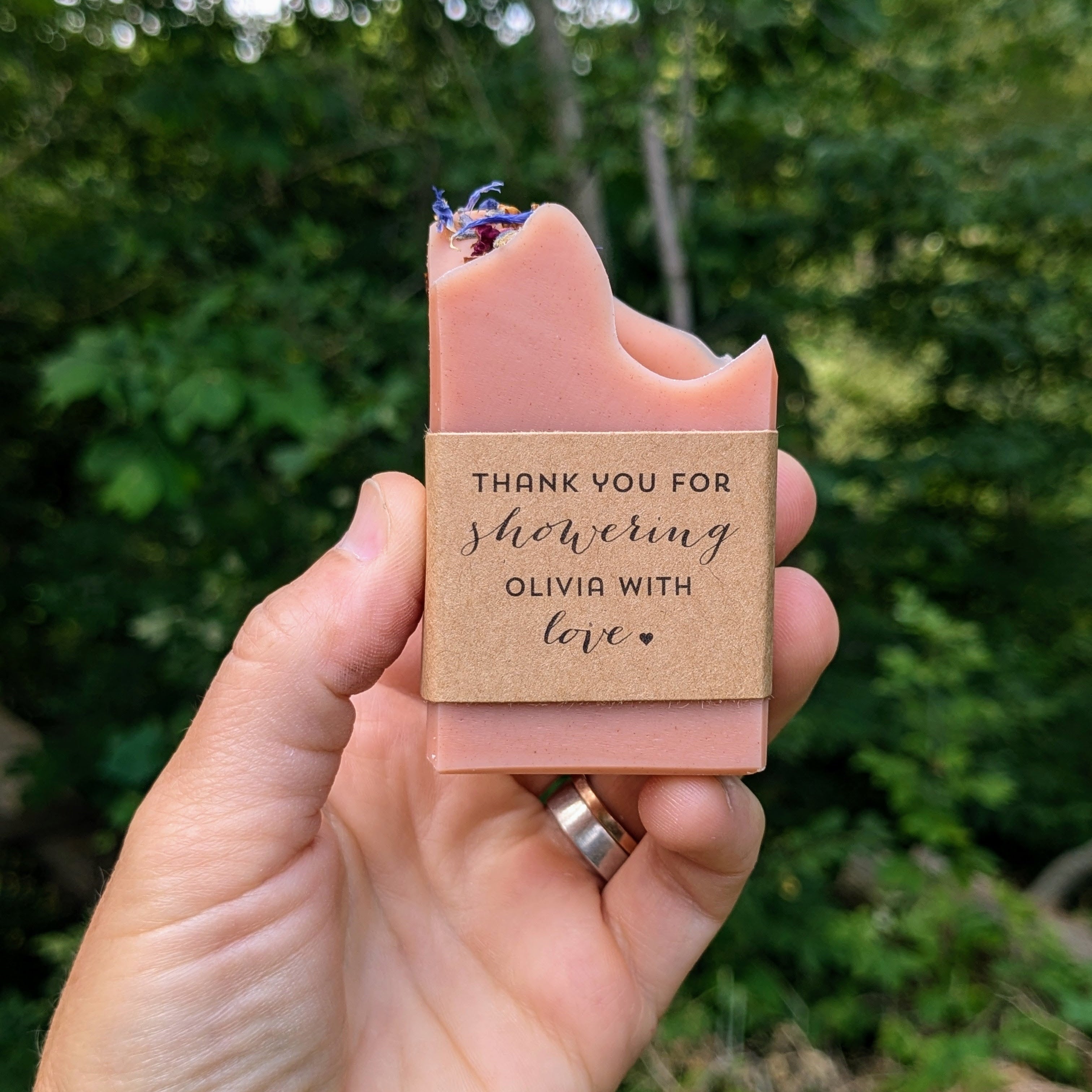 A pink handmade soap bar with custom labels reading "thank you for showering Olivia with love" displayed in an outdoor setting