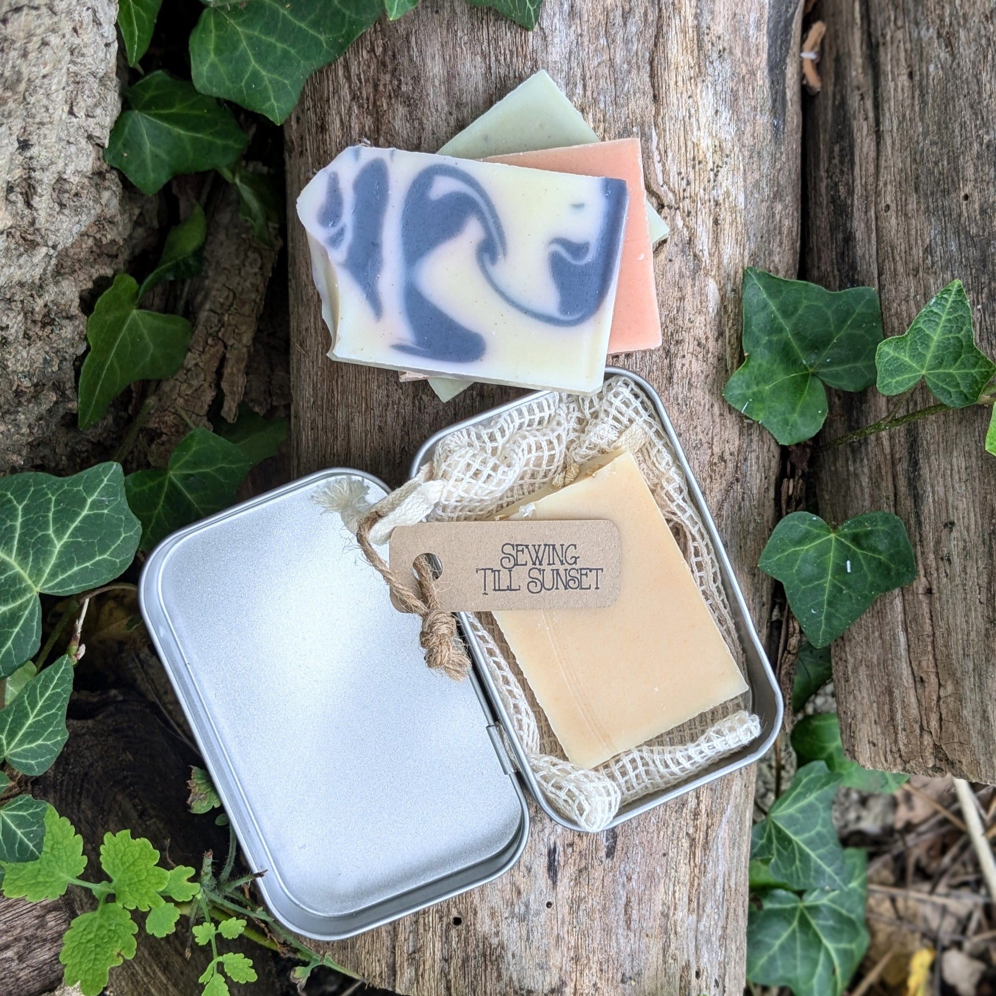 A travel tin containing a small bar of organic soap and a crocheted soap saver bag, placed on a wooden surface with green leaves and a tag that reads 'Sewing Till Sunset'.