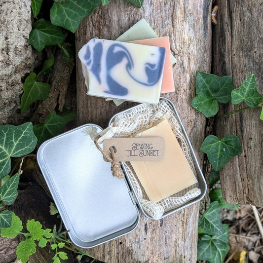A travel tin containing a small bar of organic soap and a crocheted soap saver bag, placed on a wooden surface with green leaves and a tag that reads 'Sewing Till Sunset'.