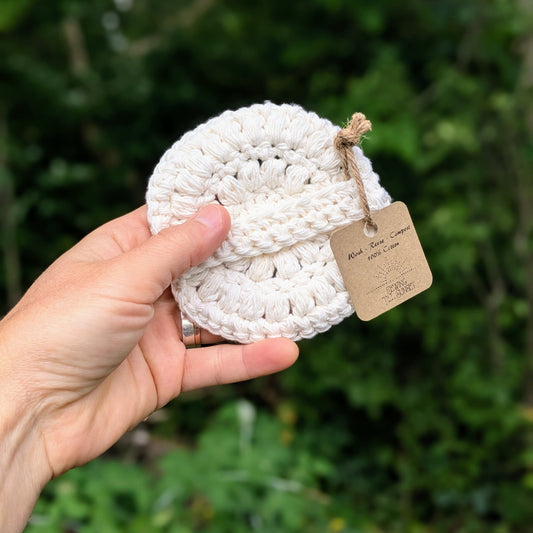 A hand holding a white crocheted facial scrubby with a strap, placed against a natural background.