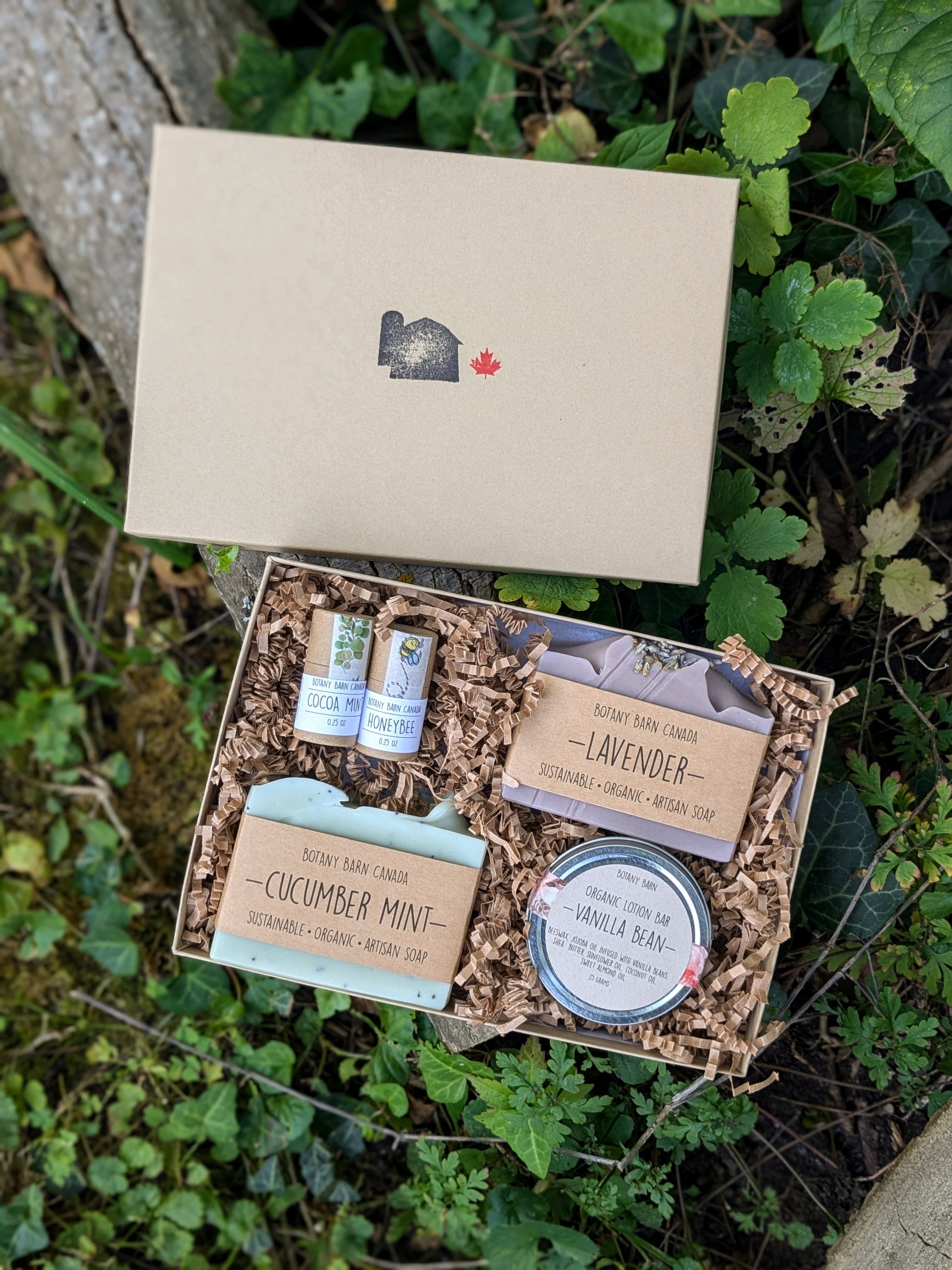 A custom gift box from Botany Barn Canada containing artisan soaps, lip balm, and a lotion bar, displayed outdoors with visible product labels and a kraft paper box.