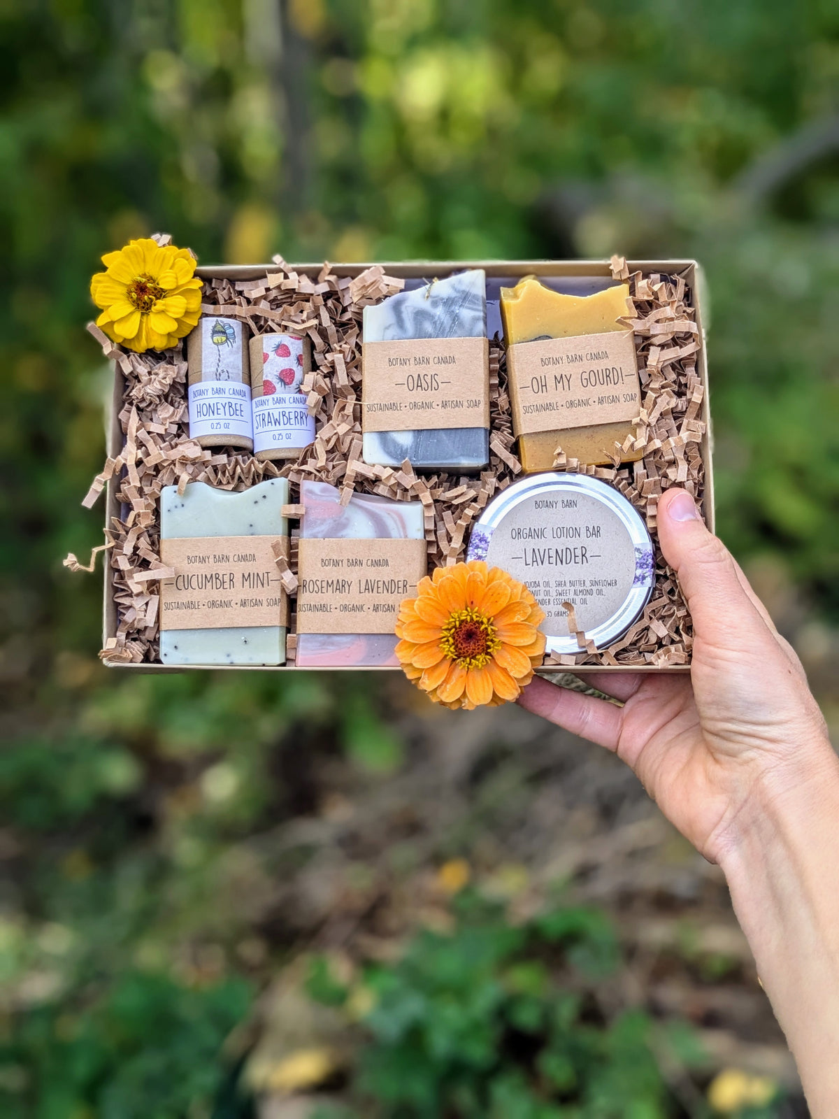 A gift set of handmade soaps, organic lotion in a metal tin, and eco-friendly lip balm tubes, all packaged in a kraft gift box with decorative flowers on top.