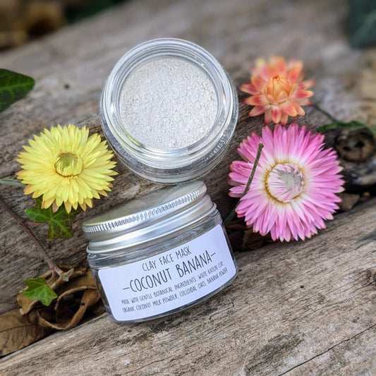 A clear glass jar containing a clay face mask with a label reading 'Coconut Banana Clay Face Mask'. The jar is surrounded by decorative flowers and leaves on a wooden surface.