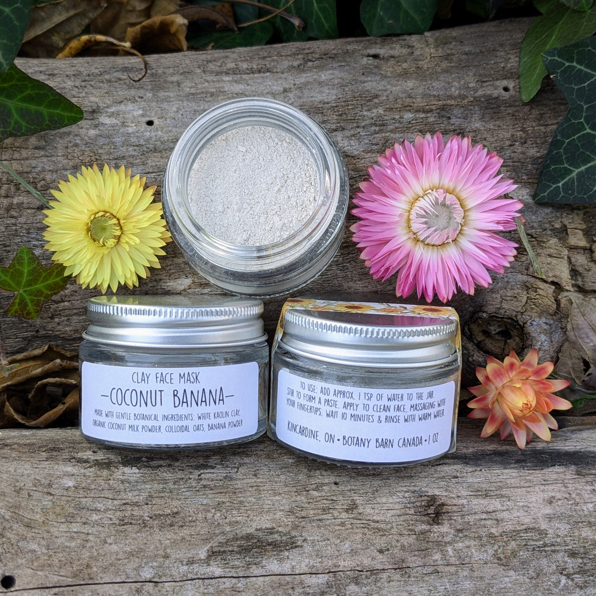 Three tins of coconut banana clay face mask on a wooden surface with flowers around.