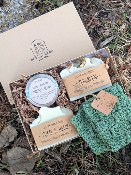An eco-friendly gift set displayed outdoors with a kraft gift box containing two soap bars, a crocheted green washcloth, and a lotion bar in a metal tin, all from Botany Barn Canada.