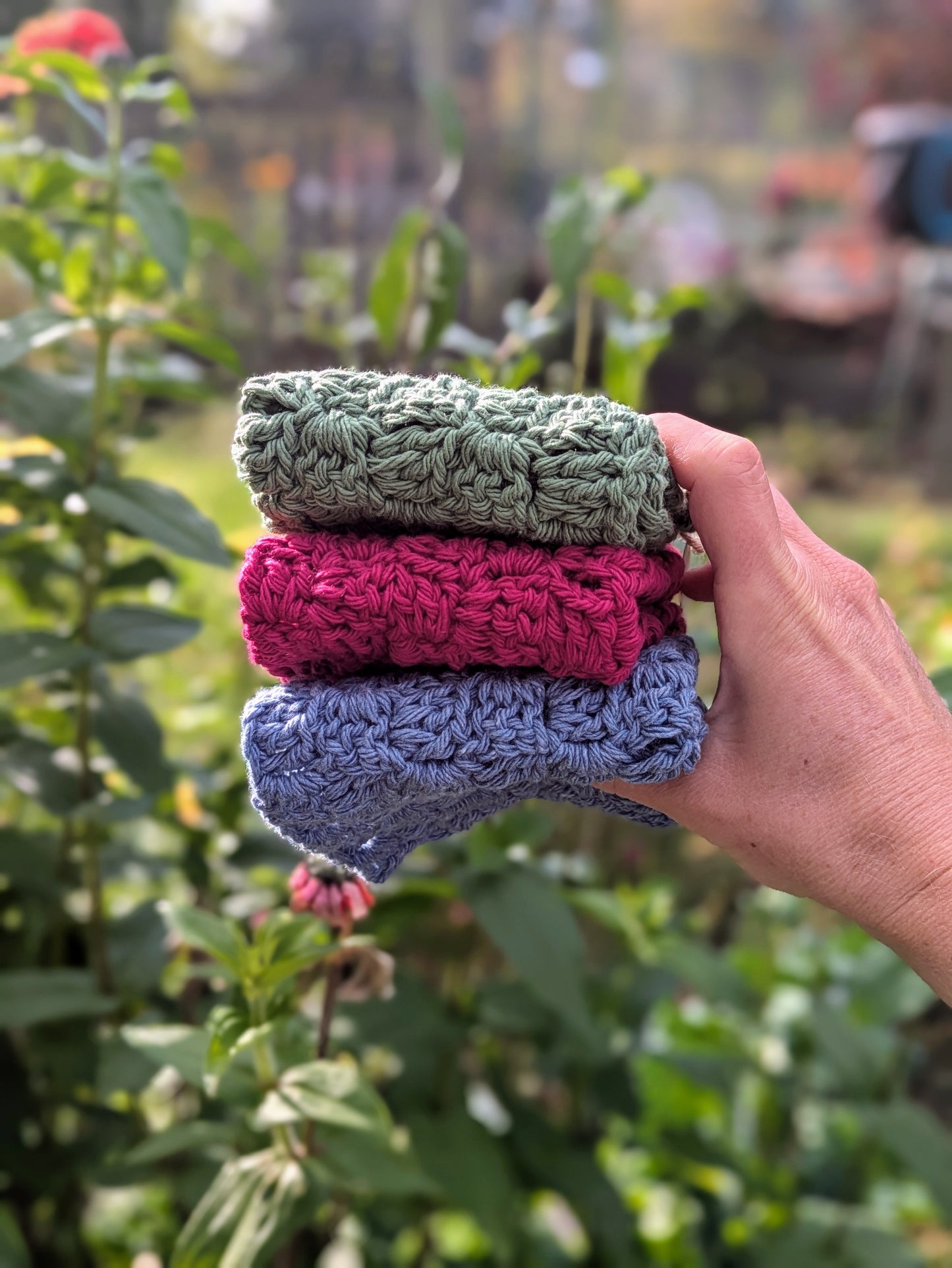 Hand holding three crocheted washcloths in front of a garden background