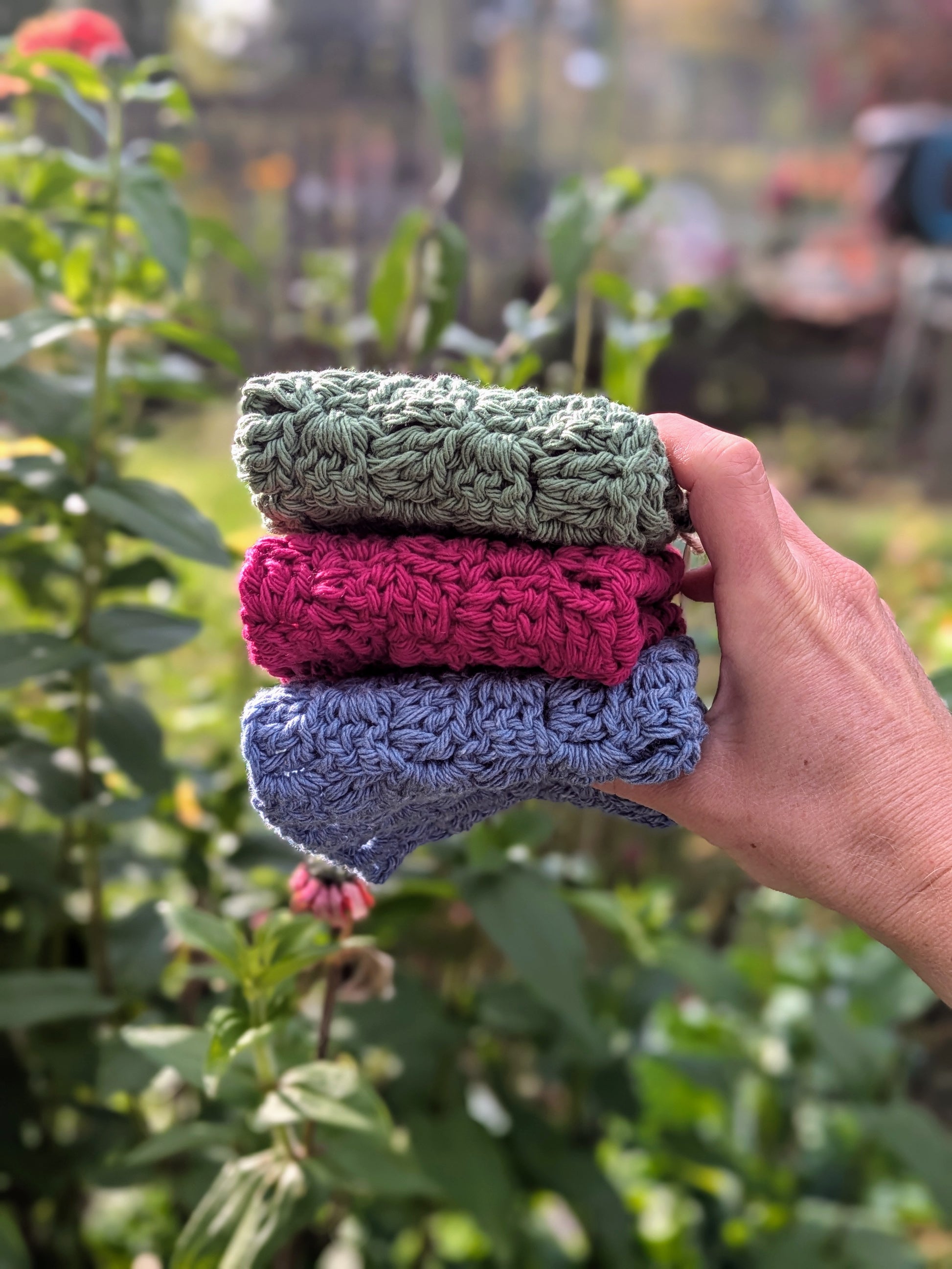 Hand holding three crocheted washcloths in front of a garden background