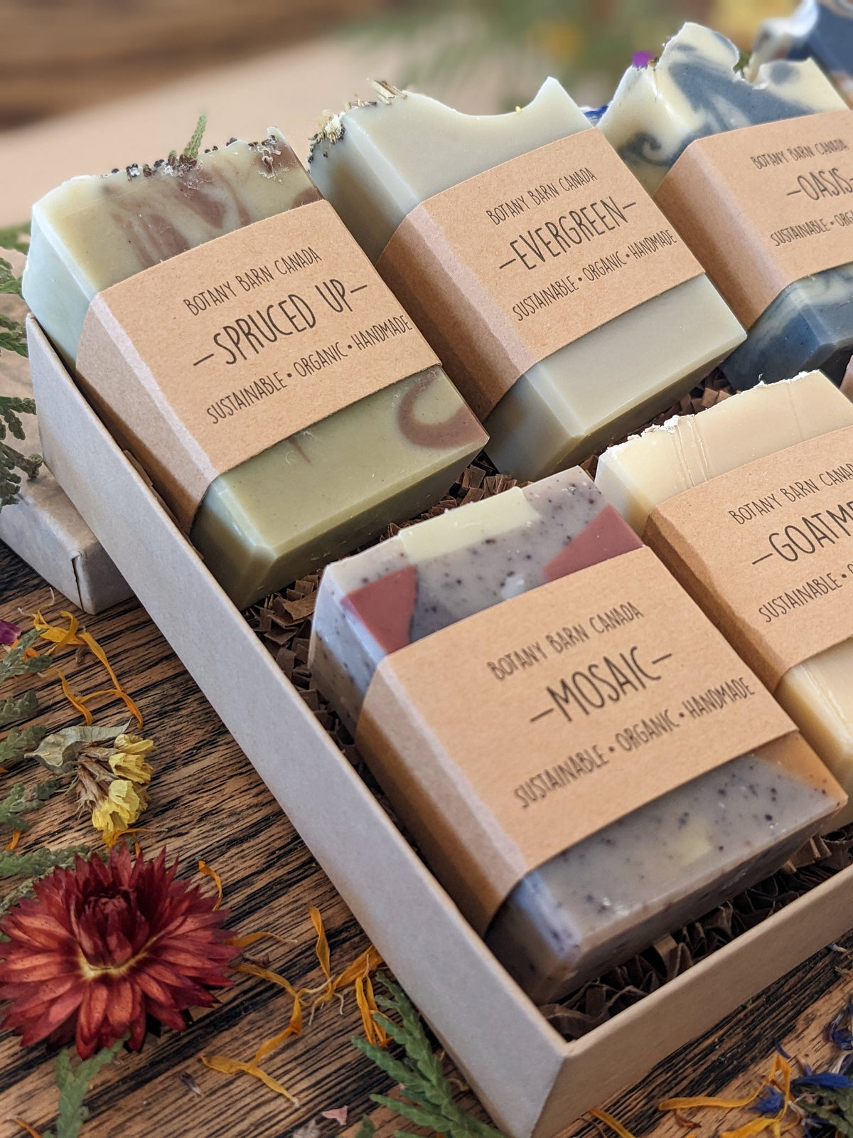 Assorted soaps with labels in a box on a wooden surface with flowers.