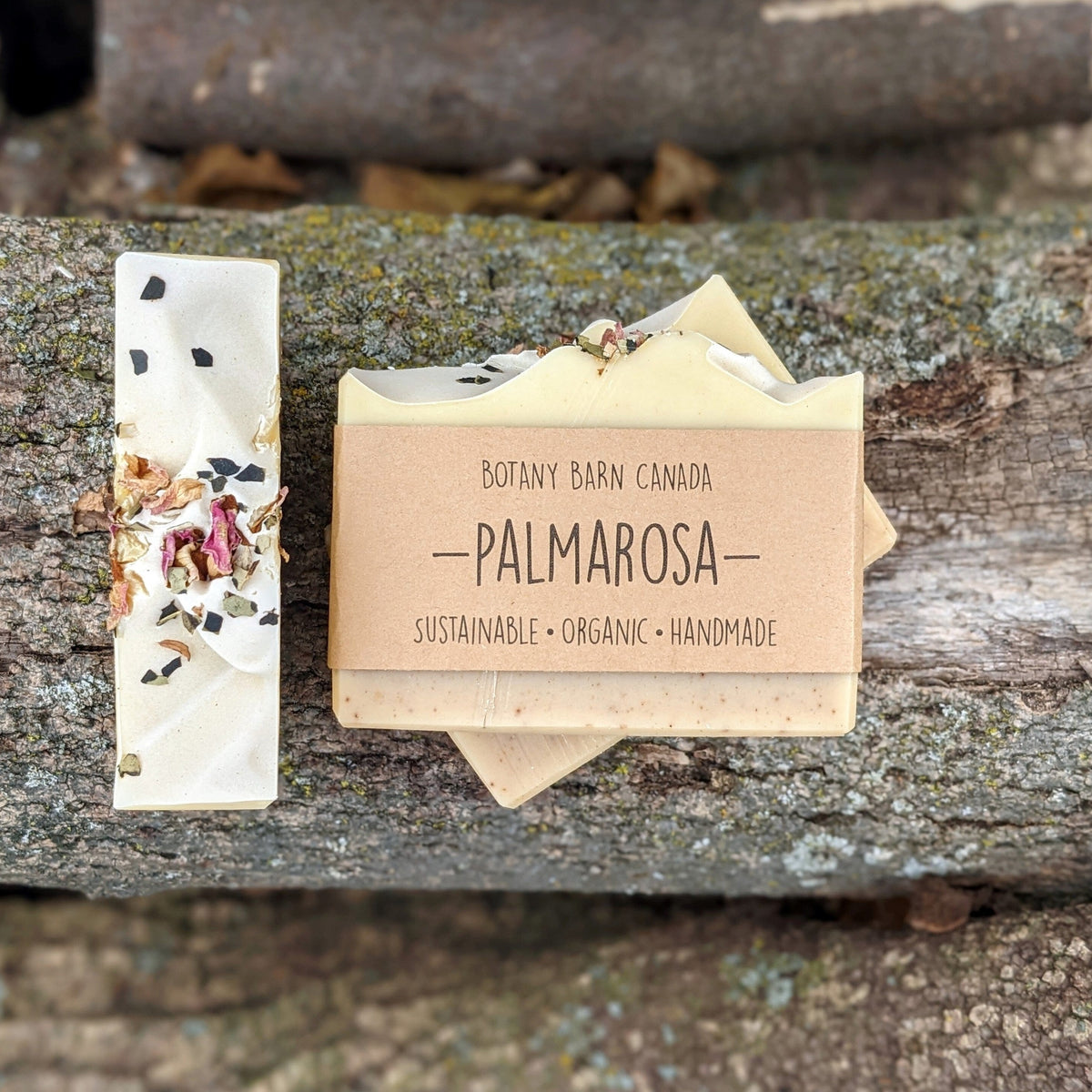 Bars of handmade artisan soap, top and front view. The soap is pale pink and yellow in colour with green and pink petals on top. A brown label reads the name of the company, Botany Barn Canada and the name of the soap, Palmarosa.
