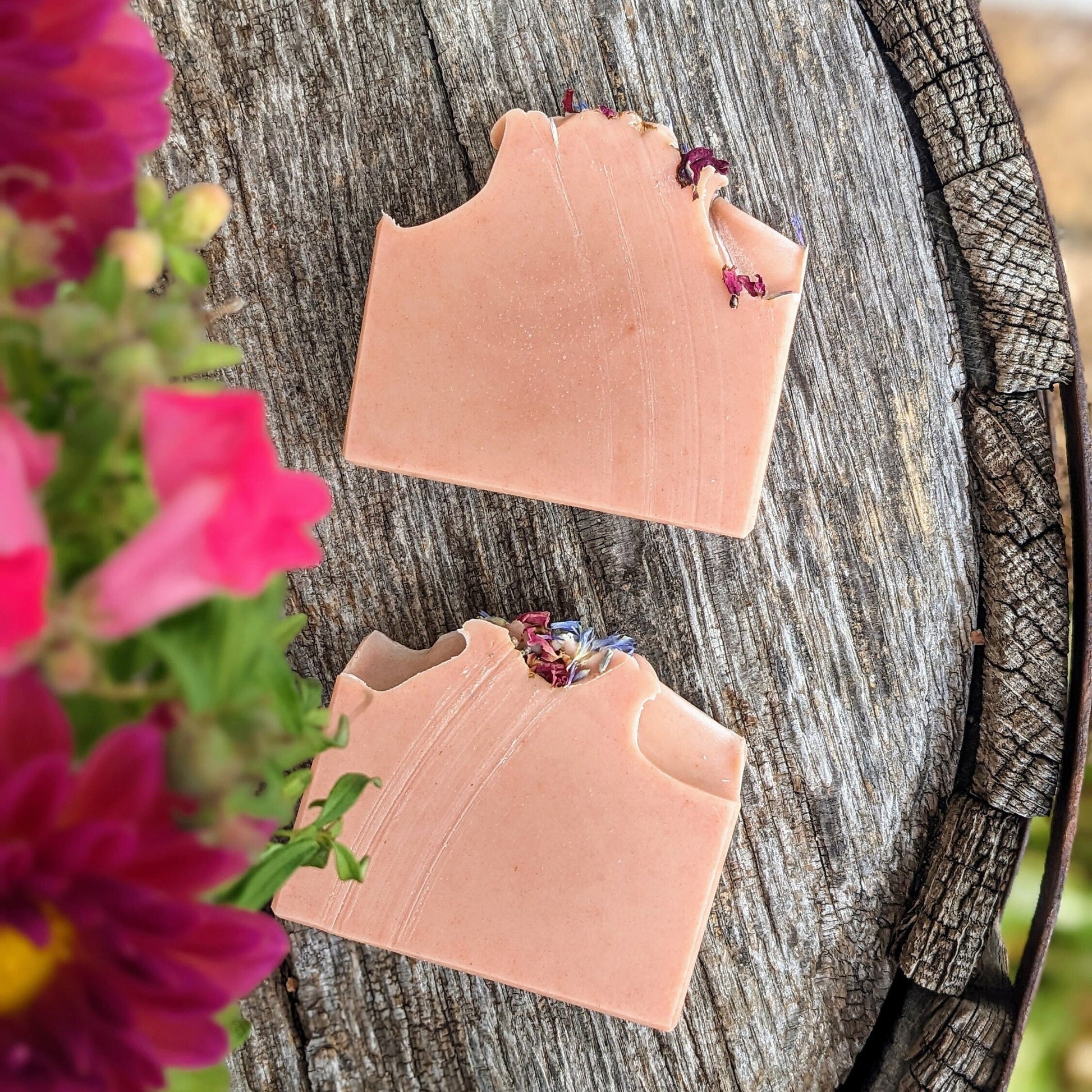 Two pink soap bars with floral elements on a wooden surface with flowers in the foreground.