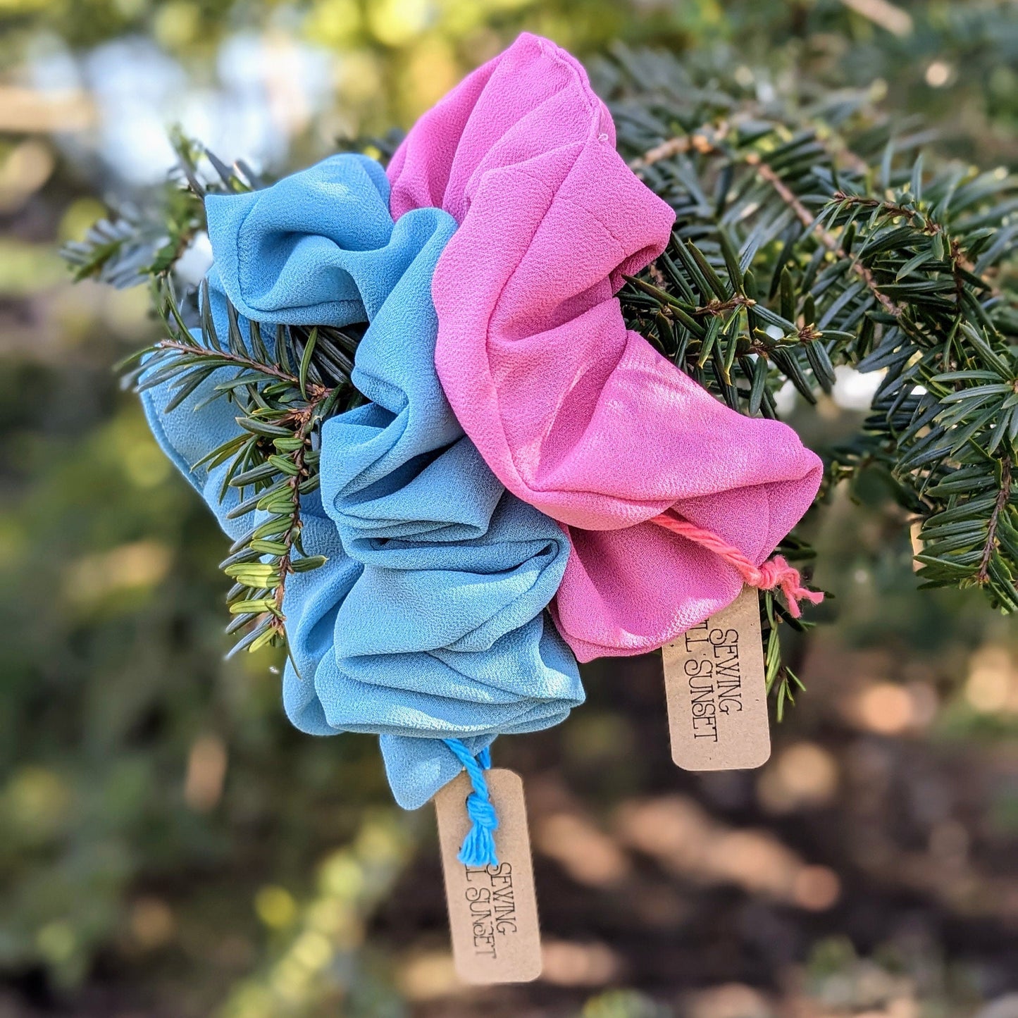 Two scrunchies, one pink and one blue, hanging on a branch with tags attached.