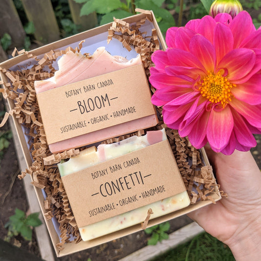 Two artisan soaps packaged in a kraft gift box, with branding visible. The box is held in a person's hand, with a pink flower partially visible in the background.