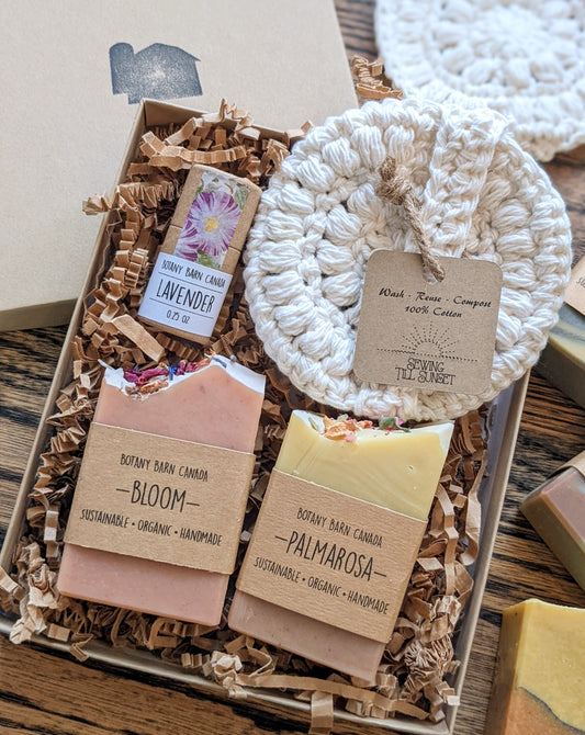 A gift set including two mini soap bars, a white crocheted face scrubby, and a lip balm in an eco-friendly tube, all packaged in a kraft gift box.