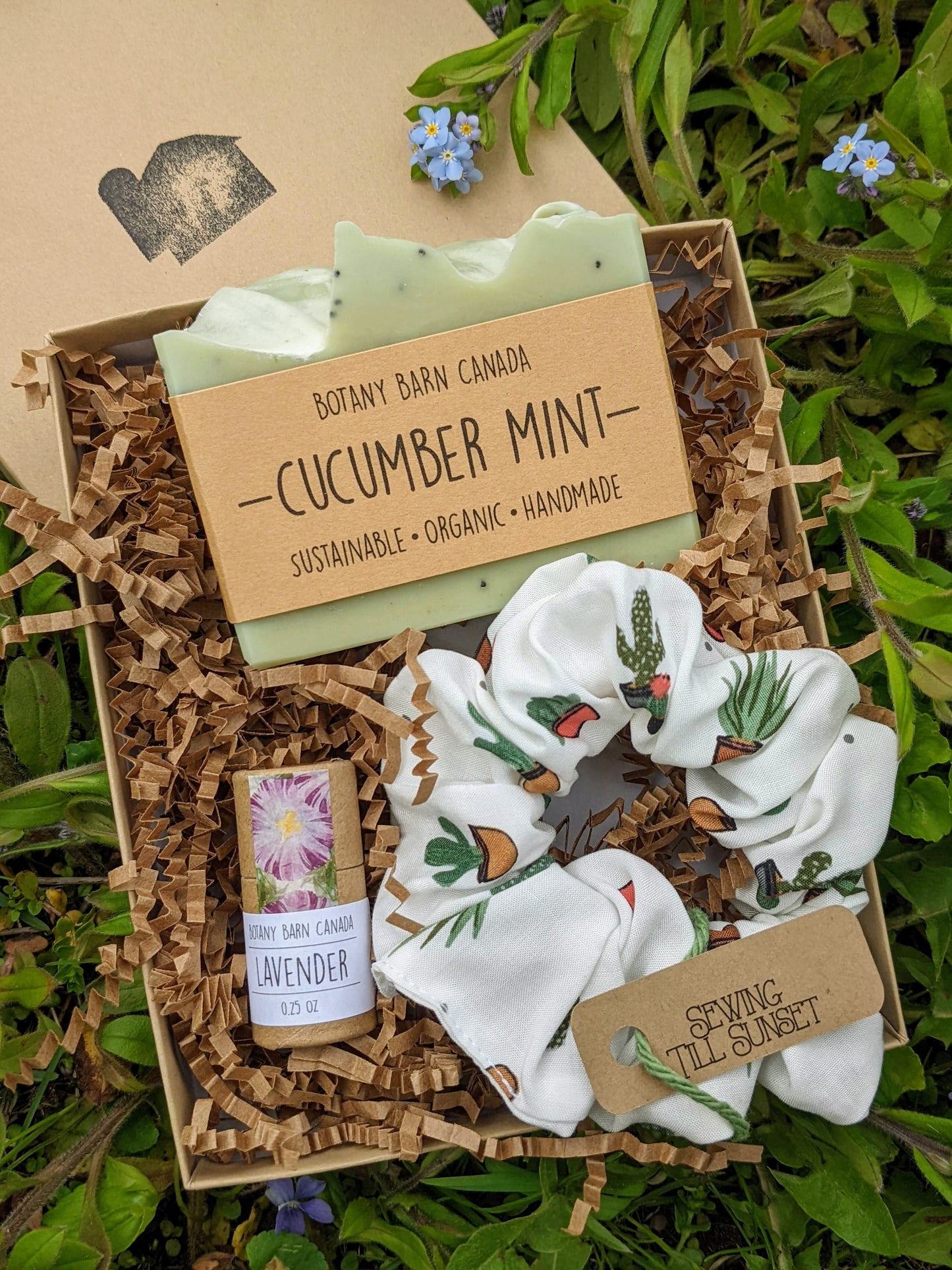 An eco-friendly gift set presented in a kraft gift box, containing a cucumber mint scented organic soap bar, a biodegradable lip balm in an eco-friendly tube, and a handmade white scrunchie with a cacti design.