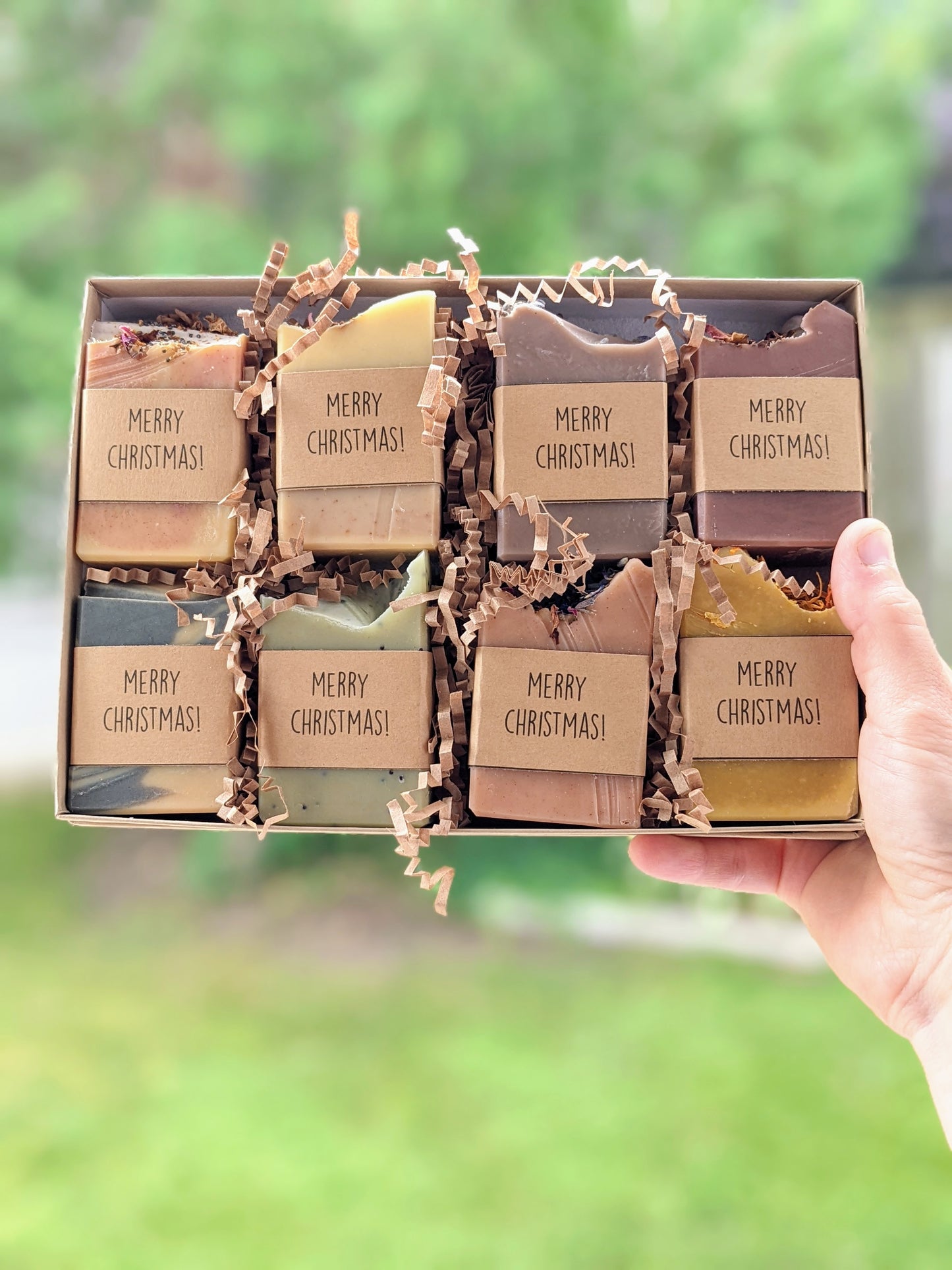 A hand holding a gift box with eight assorted half bars of soap, each with different color and labels reading Merry Christmas.
