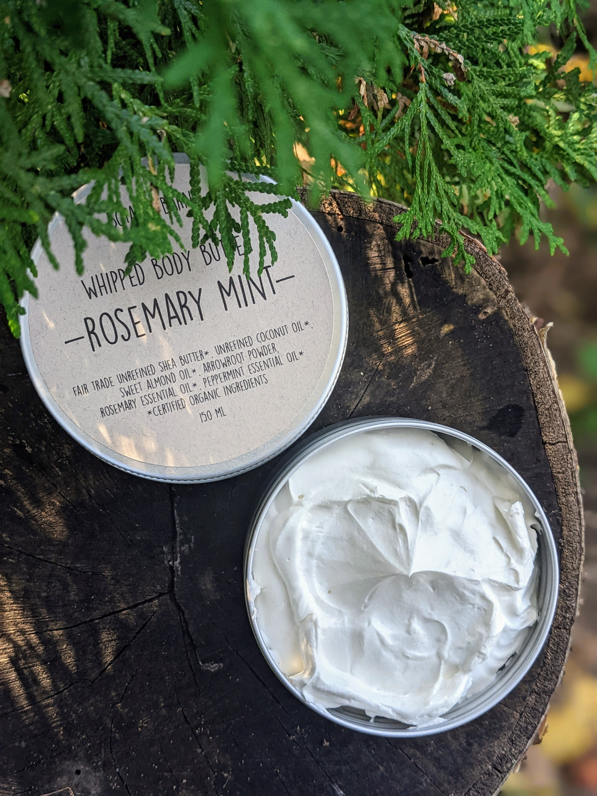 A container of whipped body butter is open, with the product inside visible as a creamy white substance. The container lid is placed beside it, "Rosemary Mint" is the scent printed on the label.