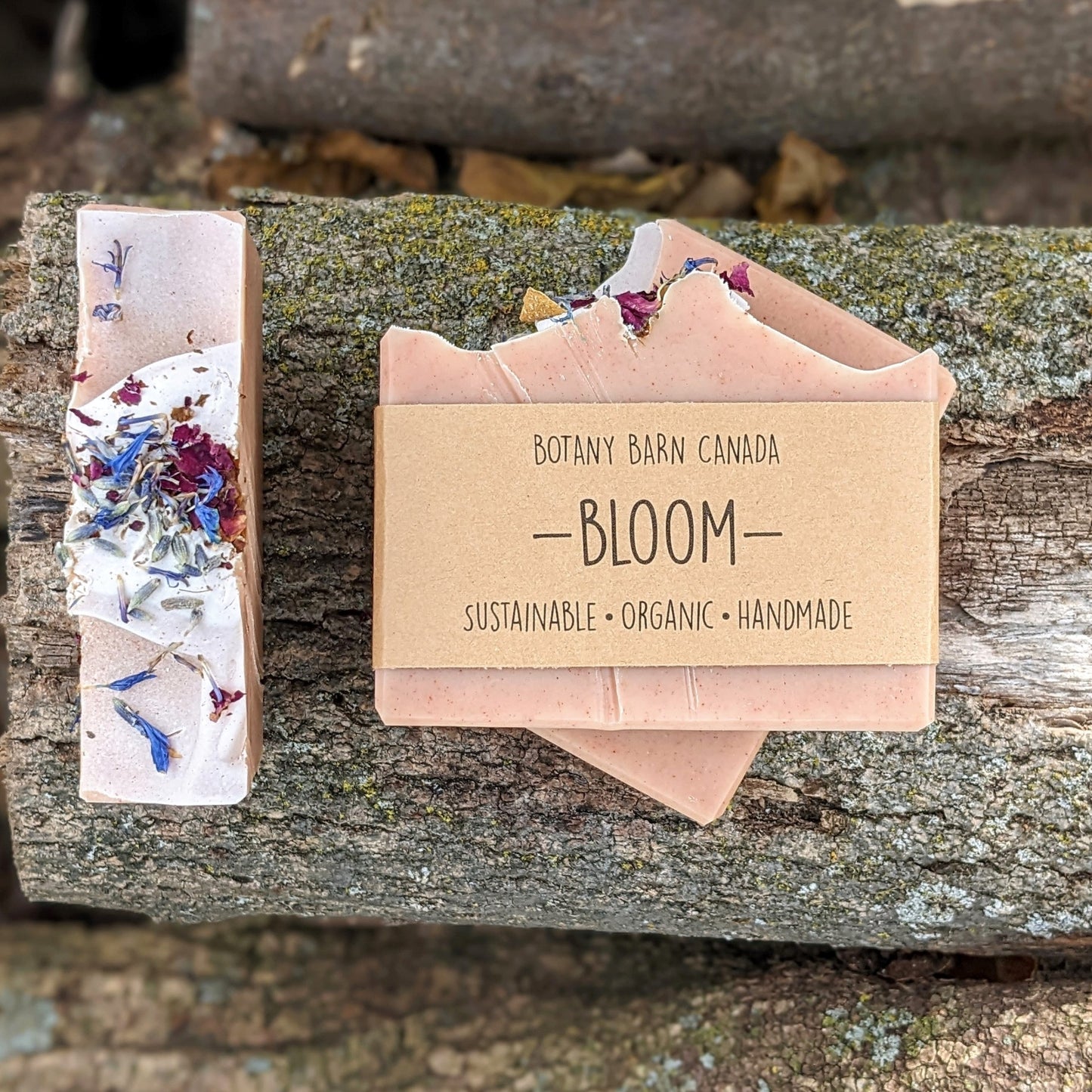 Bars of handmade artisan soap, top and front view. The soap is dusty pink in colour and decorated with brightly coloured flower petals on top. A brown label reads the name of the company, Botany Barn Canada and the name of the soap, Bloom.