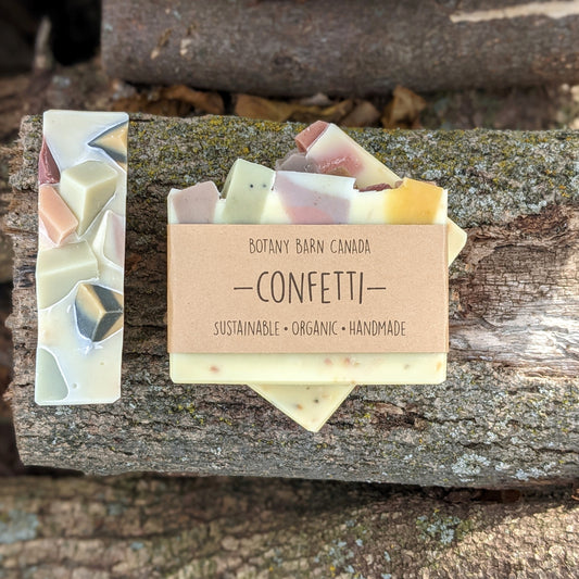 Bars of handmade artisan soap, top and front view. The soap is coloured white with specks of multicolour throughout. A brown label reads the name of the company, Botany Barn Canada and the name of the soap, Confetti.