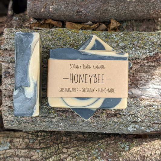 Bars of handmade artisan soap, top and front view. The soap is layered yellow and black in colour, designed to look like a bee. A brown label reads the name of the company, Botany Barn Canada and the name of the soap, Honeybee.