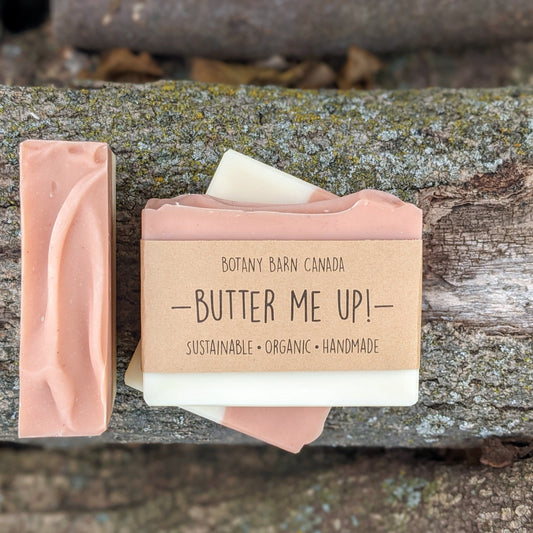 Bars of handmade artisan soap, top and front view. The soap is pink and white in colour. A brown label reads the name of the company, Botany Barn Canada and the name of the soap, Butter Me Up! Company values are listed: sustainable, organic, handmade