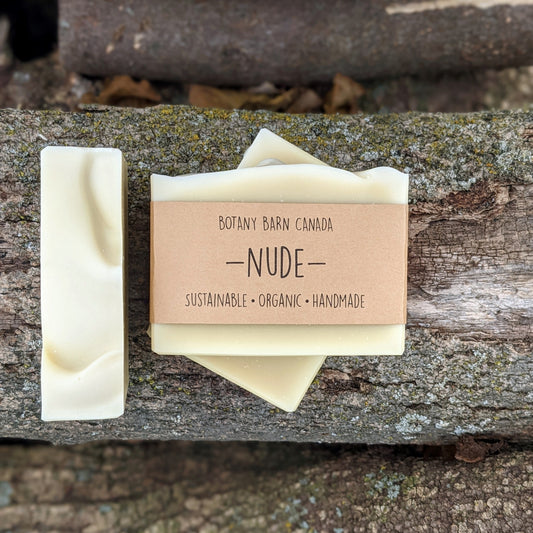 Bars of handmade artisan soap, top and front view. A simple bar of soap, white in colour. A brown label reads the name of the company, Botany Barn Canada and the name of the soap, Nude. Three company values are listed: sustainable, organic, handmade.