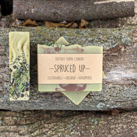 Bars of handmade artisan soap, top & front view. The soap is swirled green and brown in colour. The soap is decoratively topped with sprigs of cedar. A brown label reads the name of the company, Botany Barn Canada and the name of the soap, Spruced Up