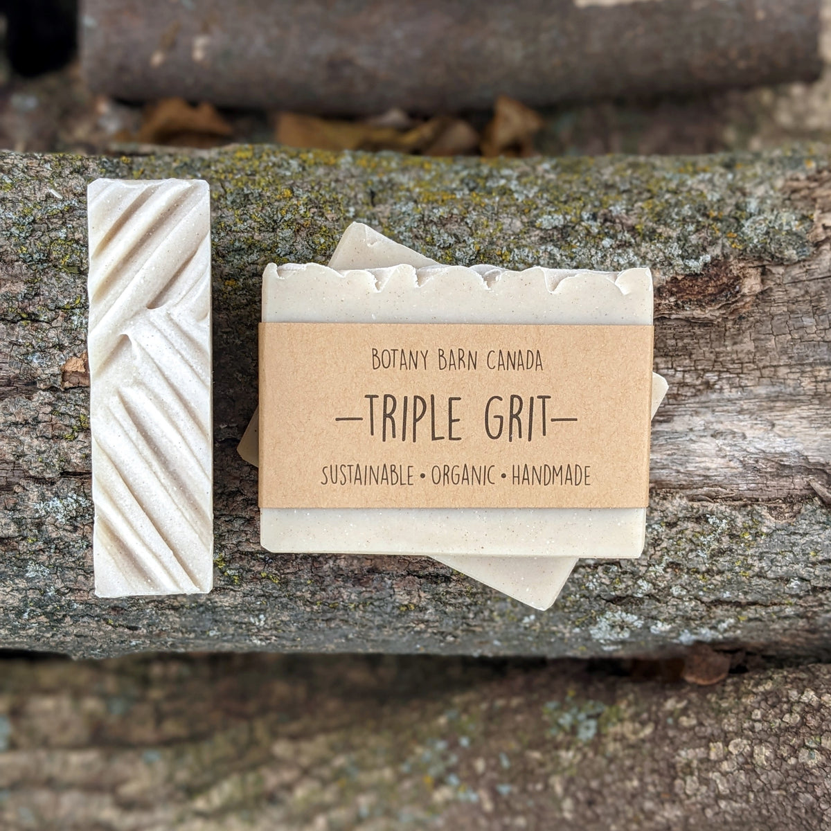 Bars of handmade artisan soap, top and front view. The soap is grey in colour. A brown label reads the name of the company, Botany Barn Canada and the name of the soap, Triple Grit. Three company values are listed: sustainable, organic, handmade.