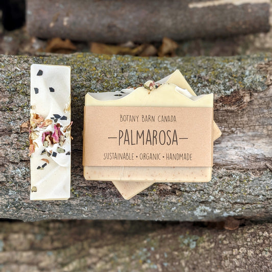 Bars of handmade artisan soap, top and front view. The soap is pale pink and yellow in colour with green and pink petals on top. A brown label reads the name of the company, Botany Barn Canada and the name of the soap, Palmarosa.