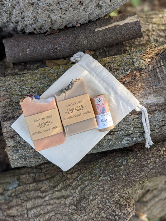 Two mini soaps and one lip balm packaged in a beige muslin bag with a drawstring, placed on a natural wood surface.