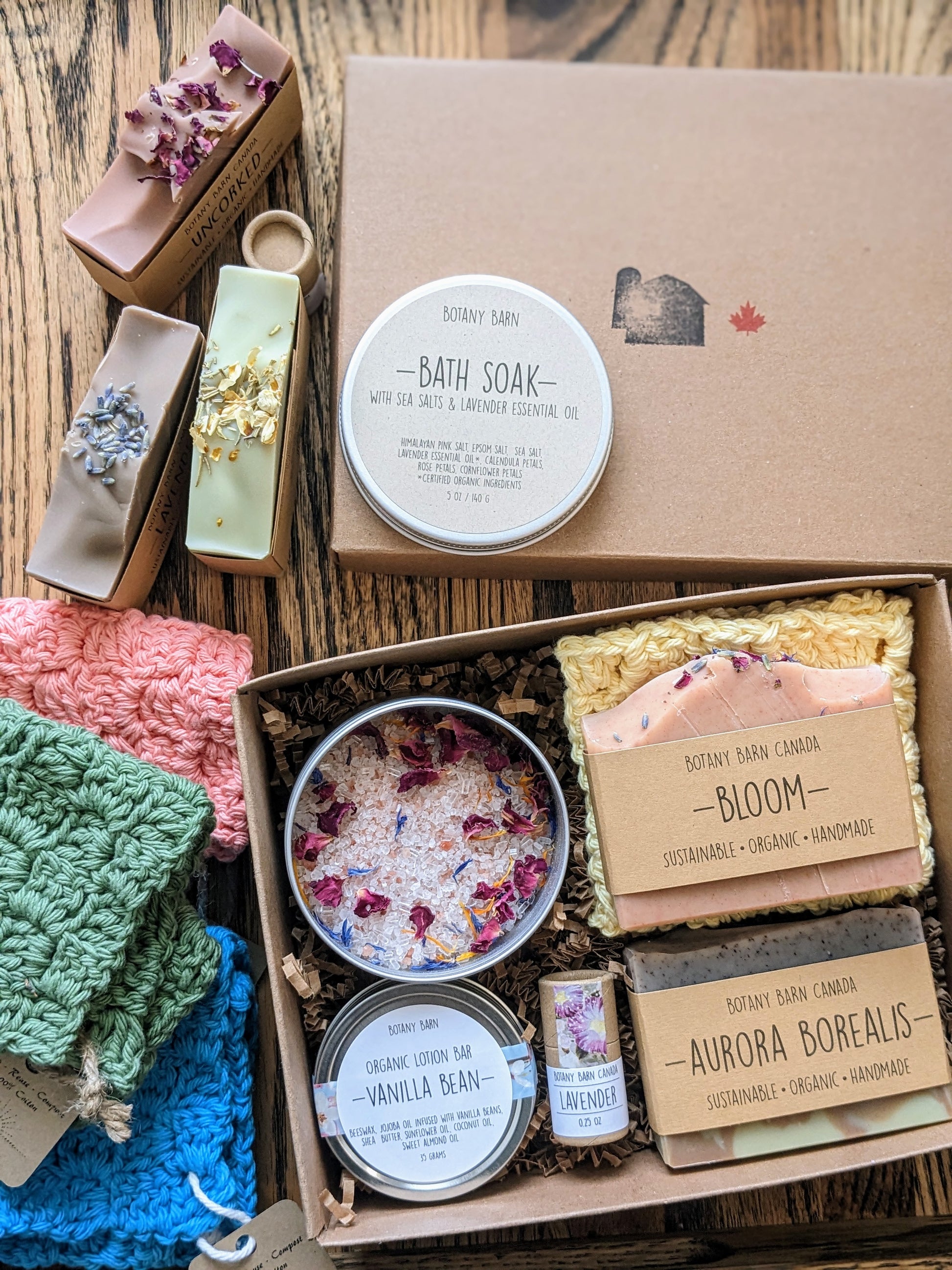 A gift box set containing various bath and body products including soap bars, a crocheted washcloth, bath soak, lotion bar, and lip balm, all displayed in an open kraft gift box.