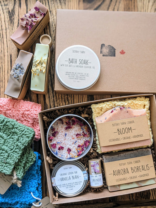 A gift box set containing various bath and body products including soap bars, a crocheted washcloth, bath soak, lotion bar, and lip balm, all displayed in an open kraft gift box.