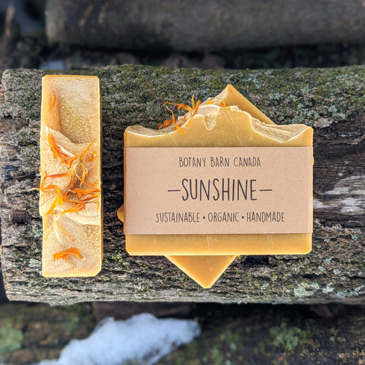 Bars of handmade artisan soap, top and front view. The soap is bright yellow in colour, with yellow flower petals on top. A brown label reads the name of the company, Botany Barn Canada and the name of the soap, Sunshine.