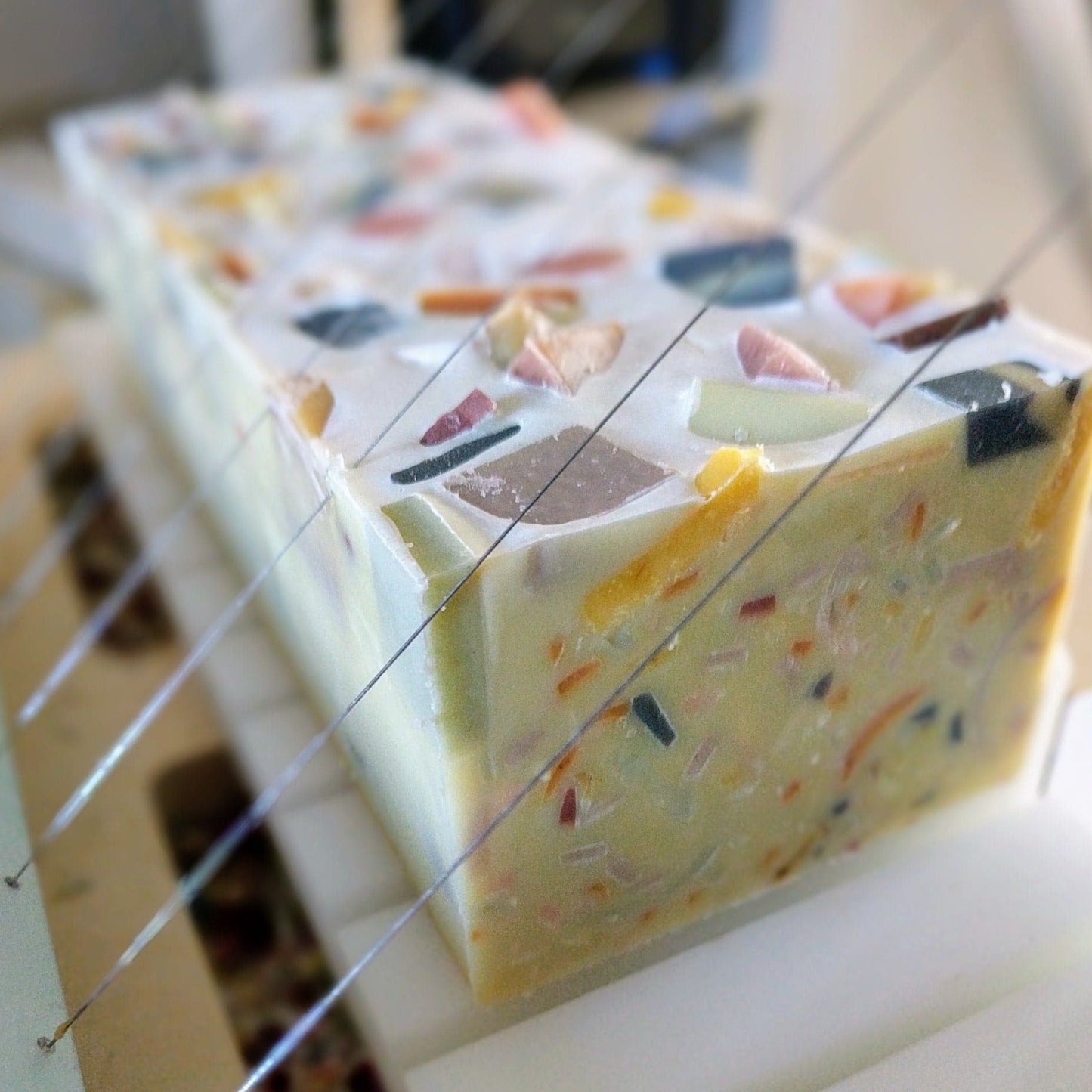 A loaf of handmade soap on a soap cutter. The soap is creamy coloured with colourful flecks throughout