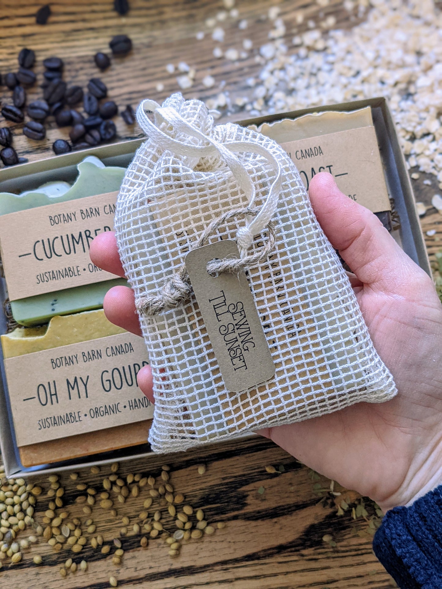 Hand holding a mesh bag with a tag over a box of organic hand soaps on a wooden surface.