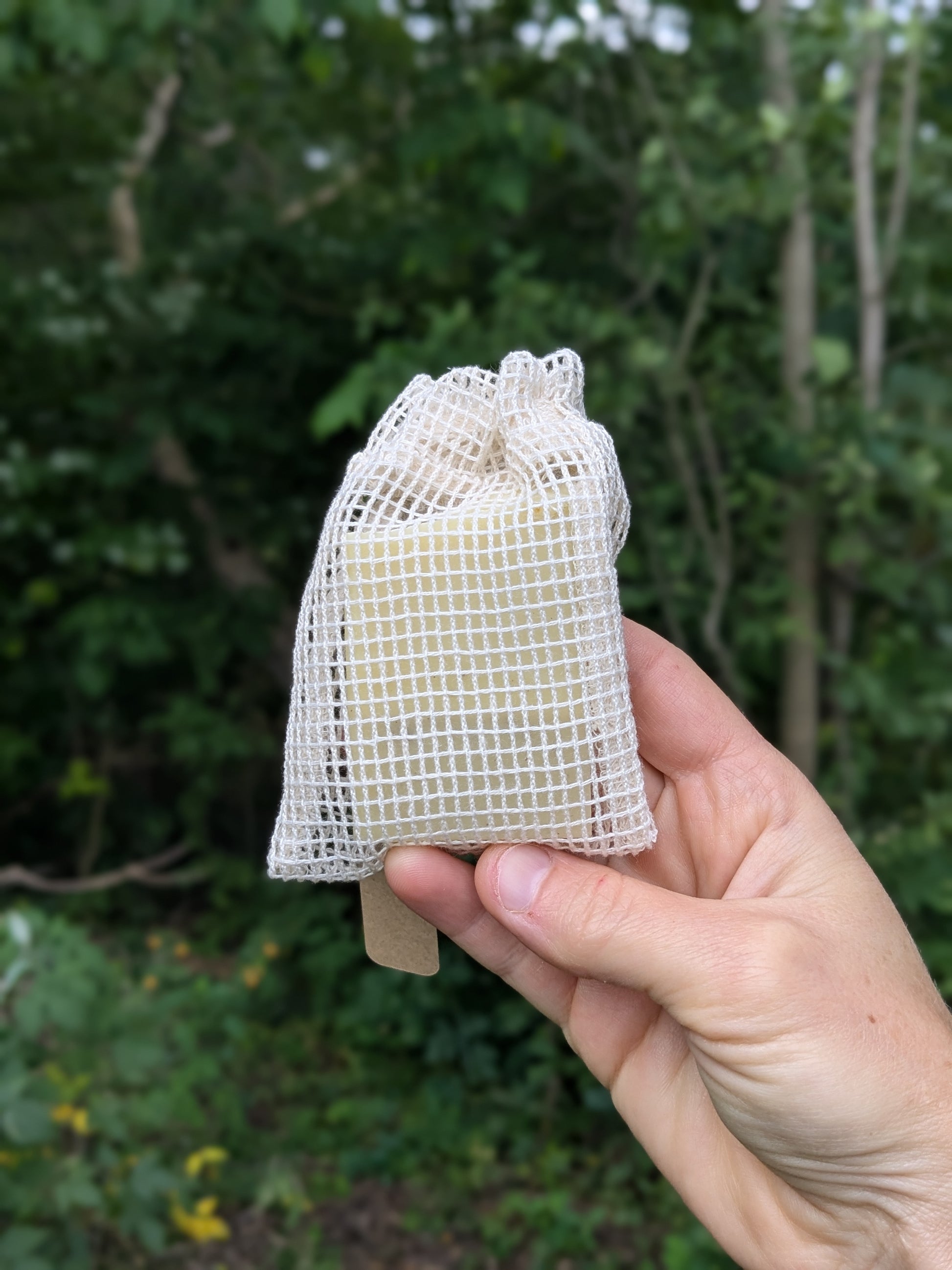 Hand holding a white mesh bag with soap inside it, against a green outdoor background