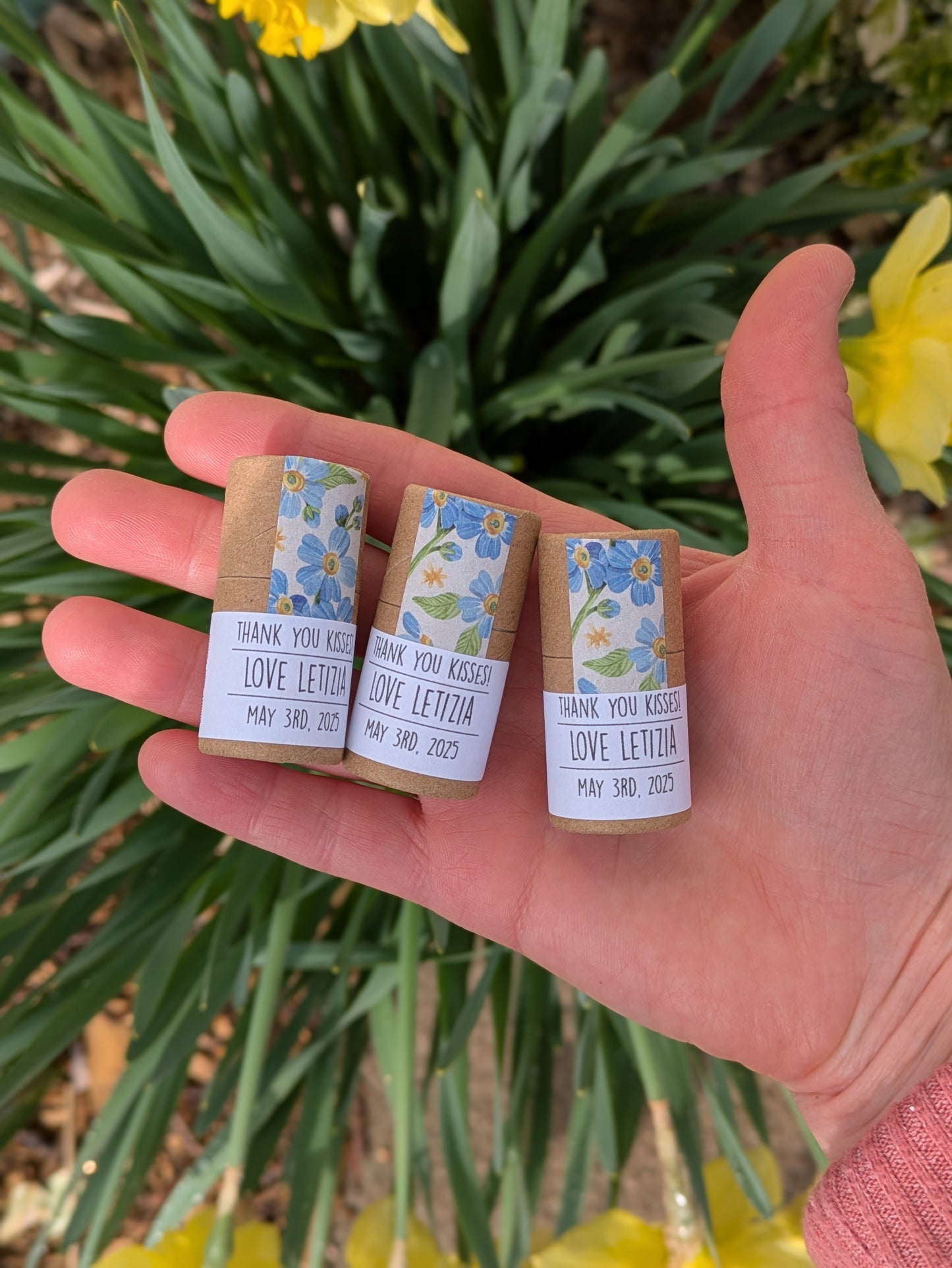Three small lip balmtubes with floral labels held in a hand, with daffodils in the background.