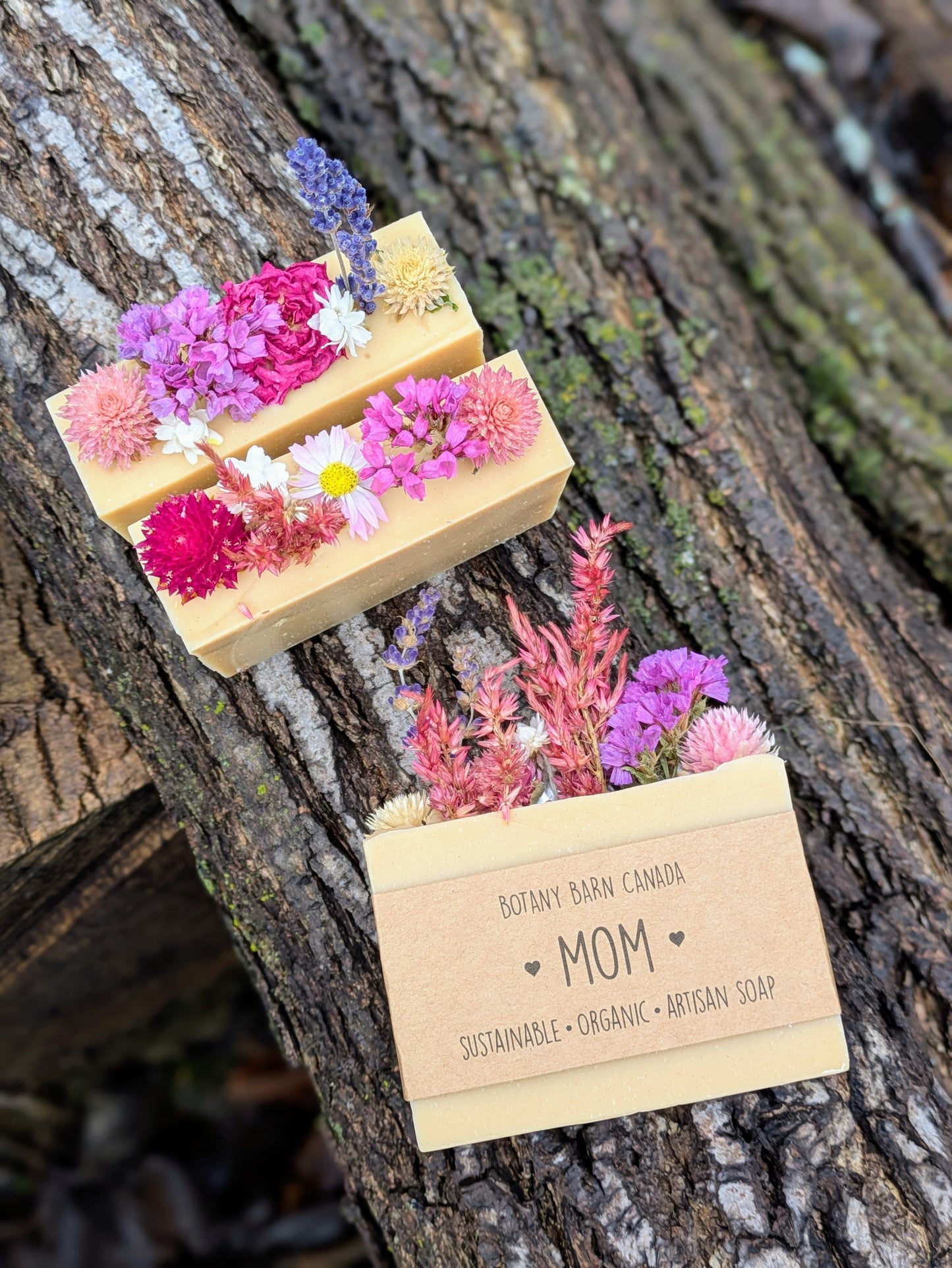 Three soap bars with floral toppings on a tree trunk, featuring a label for 'Mom' from Botany Barn Canada.
